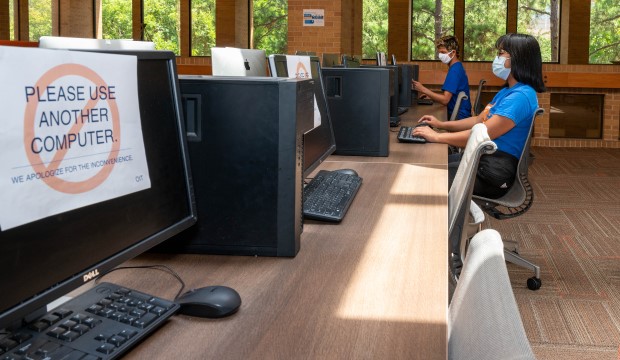 Masked students in computer lab with sign on two computers between then that says "Do Not Use This Computer."