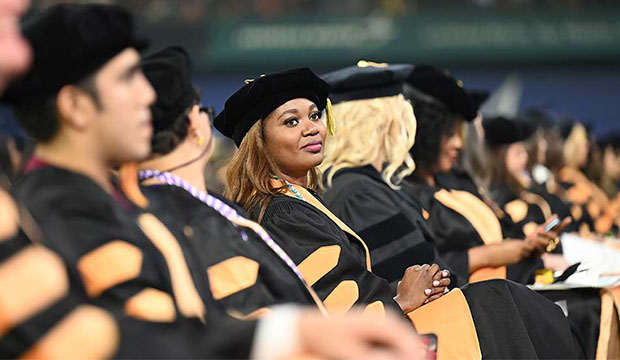 Black woman wearing doctoral graduation regalia looks to the side while sitting amongst other candidates.