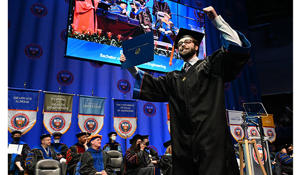 A male graduate in regalia holding up his diploma above his head.