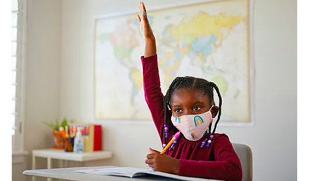 Young girl wearing a mask raising her hand in a classroom.