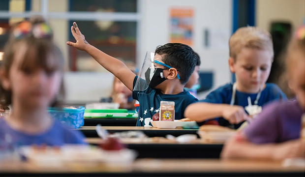 Young child in classroom wearing a mask and face shield while other children do not wear anything.