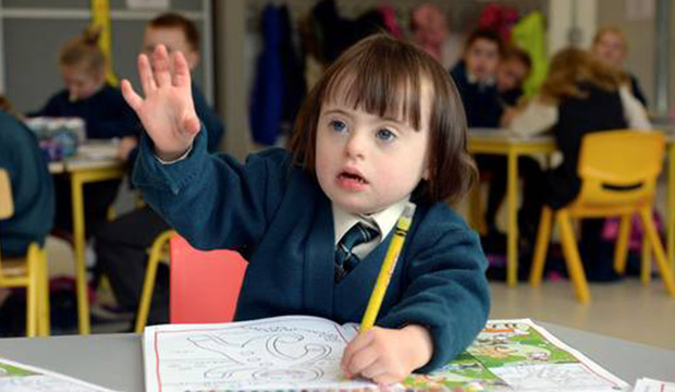 Young girl with Downs syndrome wearing school uniform and raising her hand in a classroom.