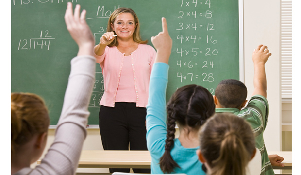 White teacher standing at blackboard, pointing to students raising their hands.