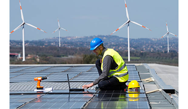 Man in hard hat examining solar panels with wind mills in background.